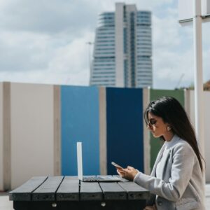 Young woman using laptop and smartphone at outdoor table. Modern cityscape background.